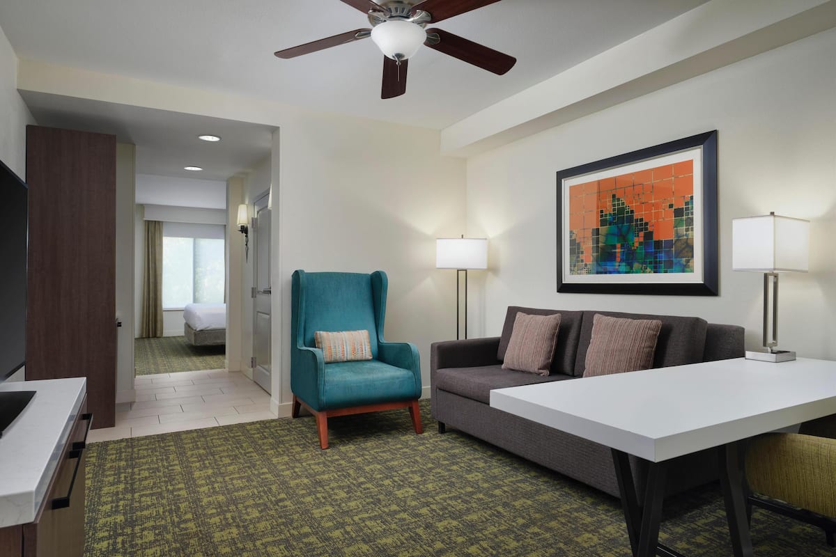 A hotel room with a teal armchair, a gray sofa, a white table, and a ceiling fan.