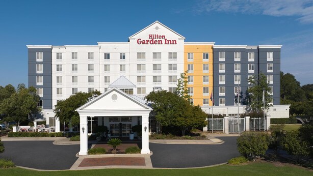 A multi-story Hilton Garden Inn with a central entrance, a covered waiting area, and a well-maintained lawn.
