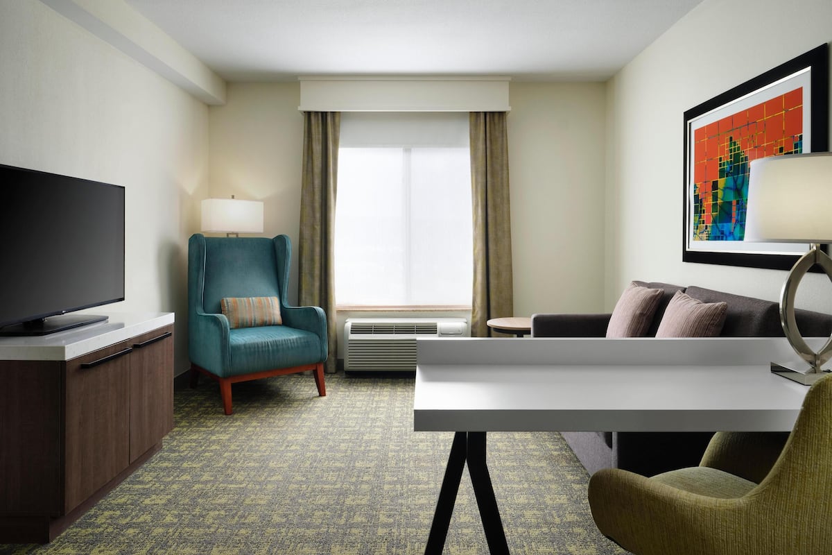 A hotel room with a flat-screen TV, a teal armchair, a white desk, a sofa, and a large window with curtains.