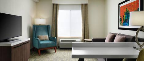 A hotel room with a flat-screen TV, a teal armchair, a white desk, a sofa, and a large window with curtains.