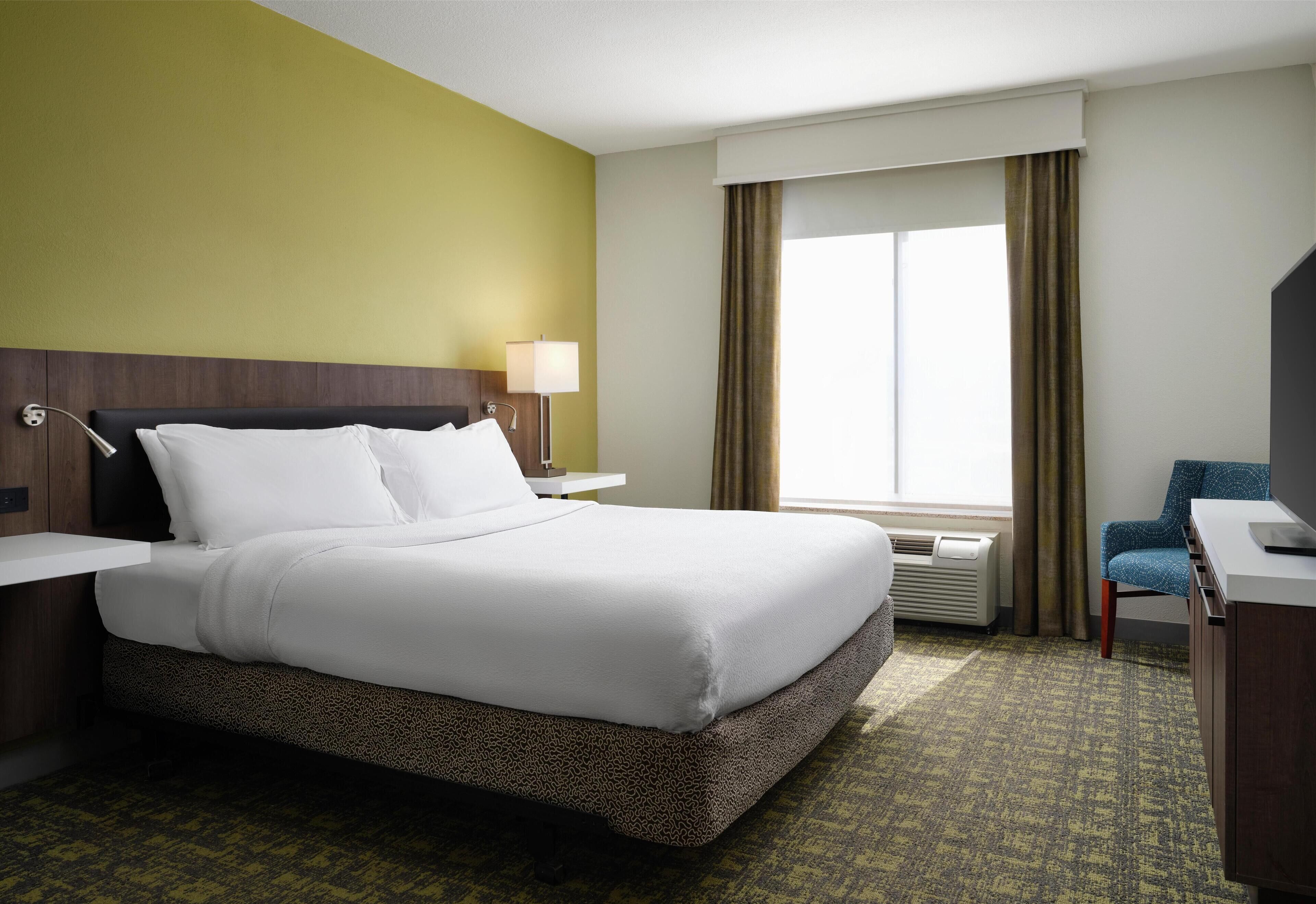 A hotel room with a large bed, a desk, a chair, a window with curtains, and a wall-mounted lamp.