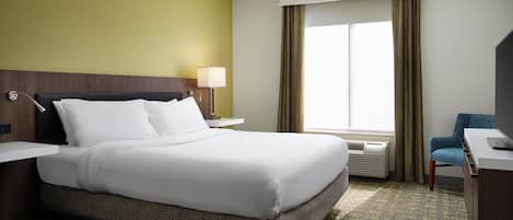A hotel room with a large bed, a desk, a chair, a window with curtains, and a wall-mounted lamp.