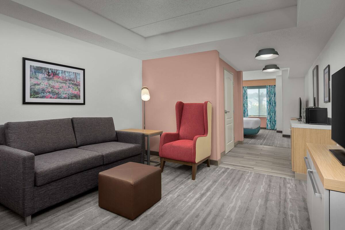 A modern hotel room with a grey sofa, a red armchair, a small table, and a TV on a wooden cabinet.