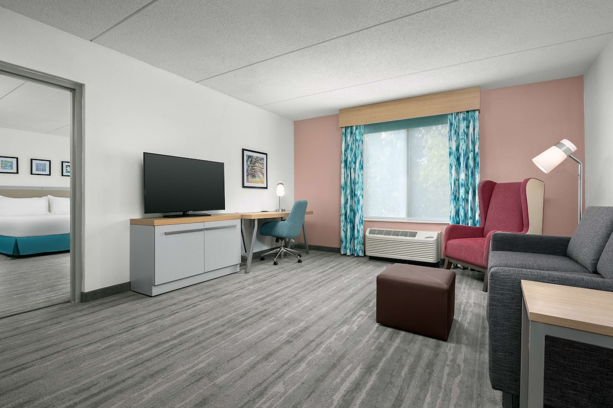 A hotel room with a bed, a desk with a television, a chair, a couch, an ottoman, and a window with curtains.