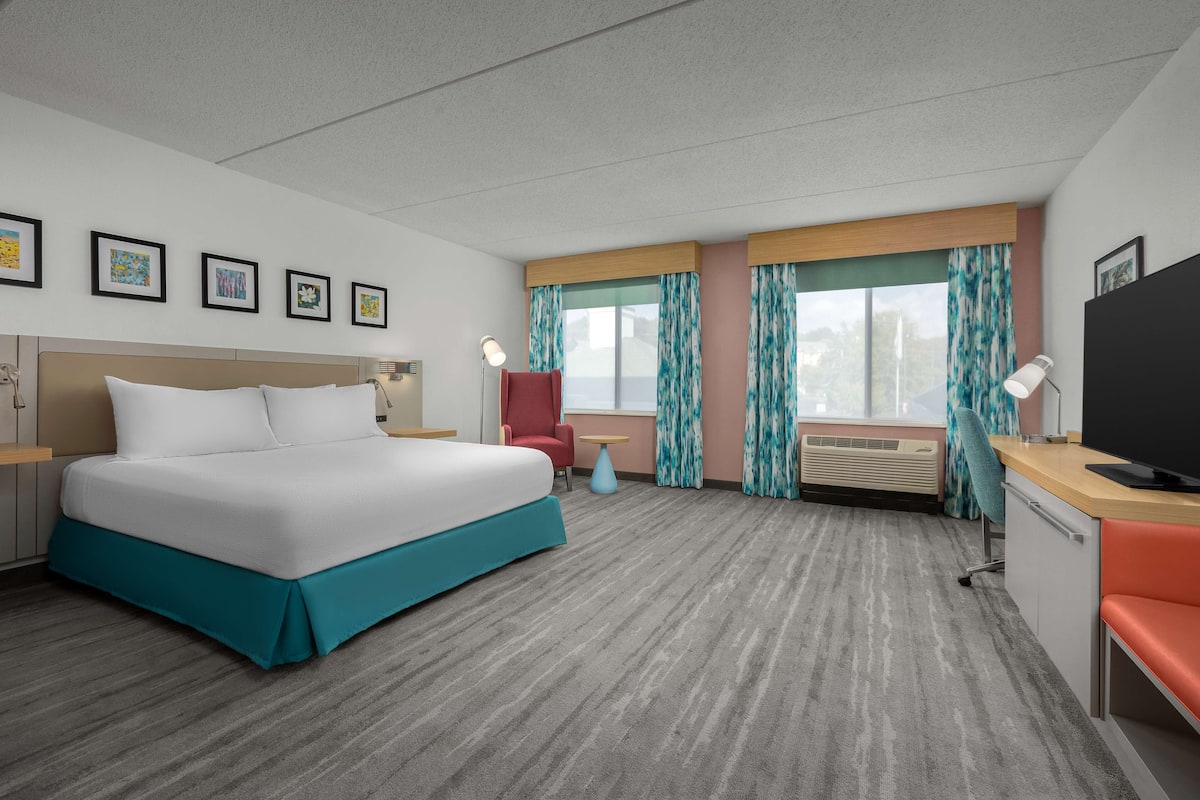 A hotel room with a large bed, a desk with a TV, a chair, a small table, and a window with curtains.