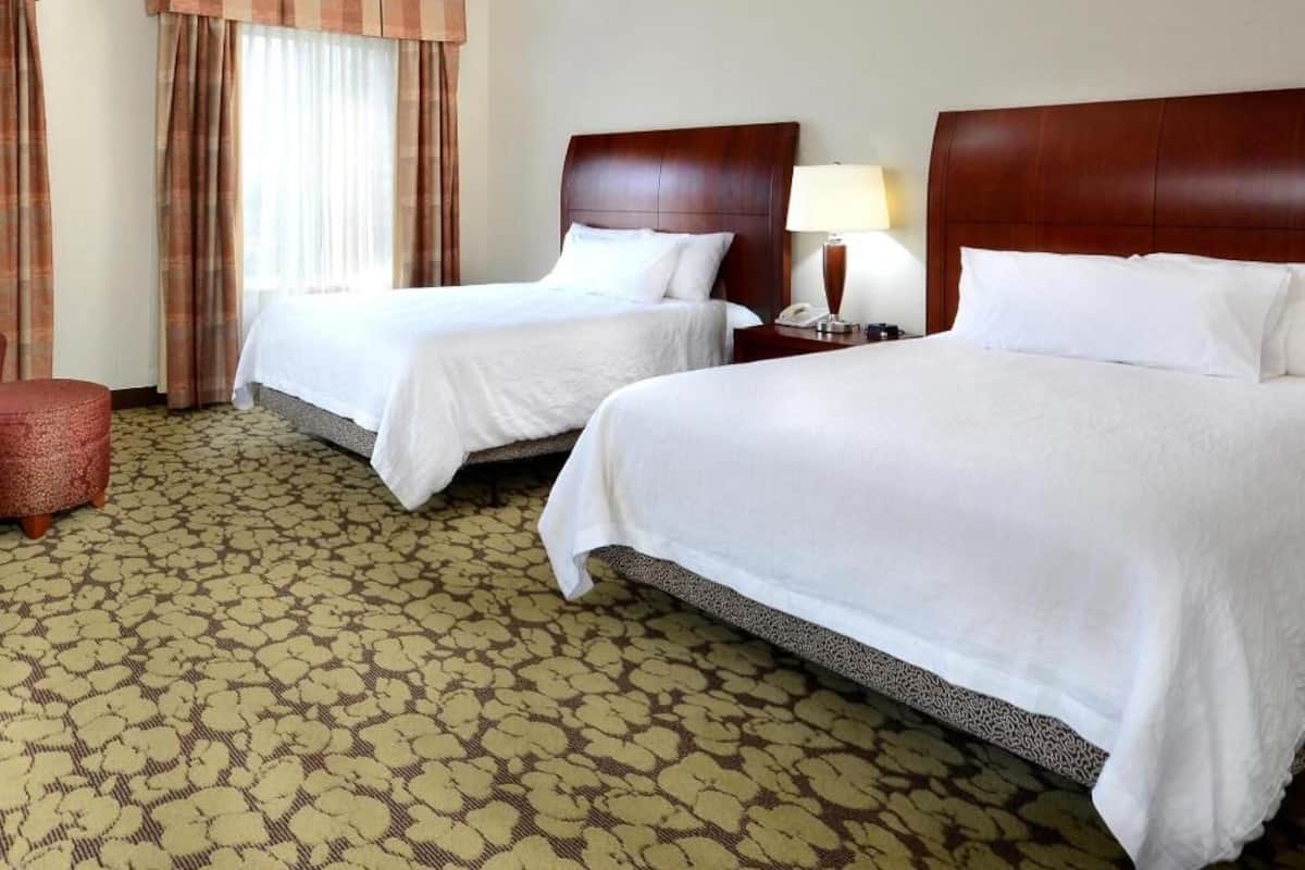 A hotel room with two beds, a patterned carpet, a red ottoman, and a window with curtains.