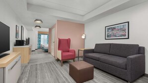 A modern hotel room with a grey sofa, a red armchair, a brown ottoman, a flat-screen TV, a coffee machine, and a framed picture on the wall. - Hilton Garden Inn Greensboro (Greensboro)