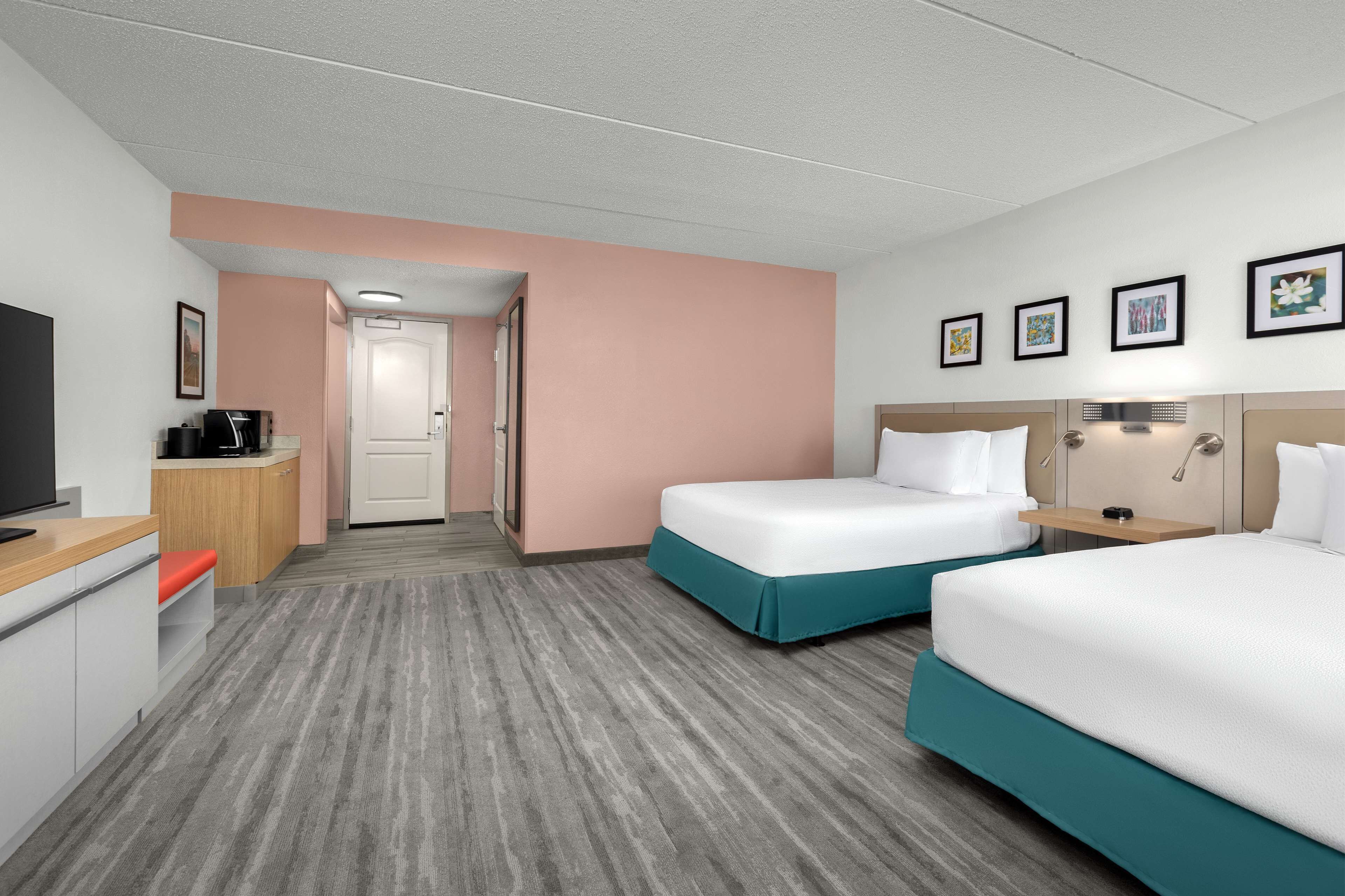 A hotel room with two beds, a desk, a TV, and a door leading to a hallway.