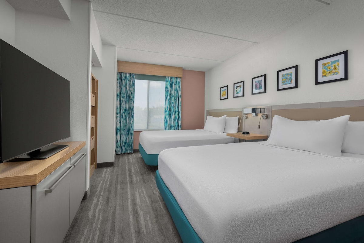 A hotel room with two beds, a flat-screen TV, a desk with a computer, and a window with curtains.