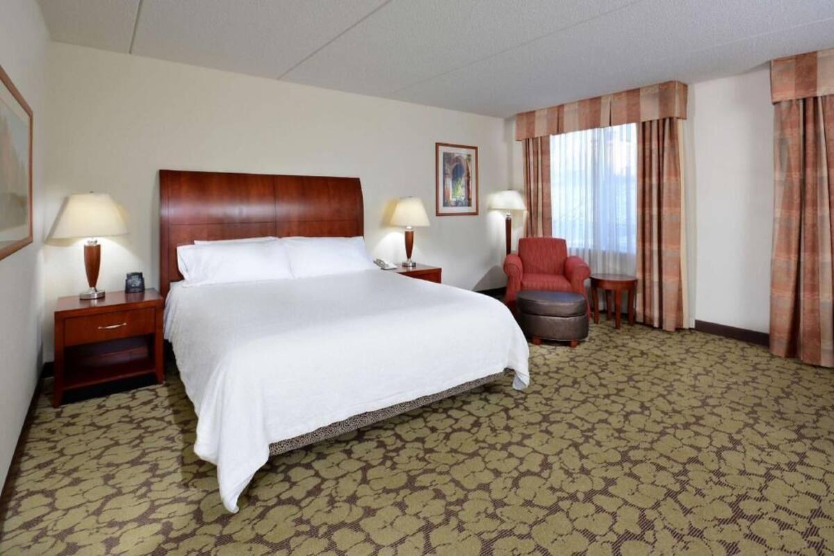 A hotel room with a large bed, two bedside tables with lamps, a red armchair, and a window with curtains.