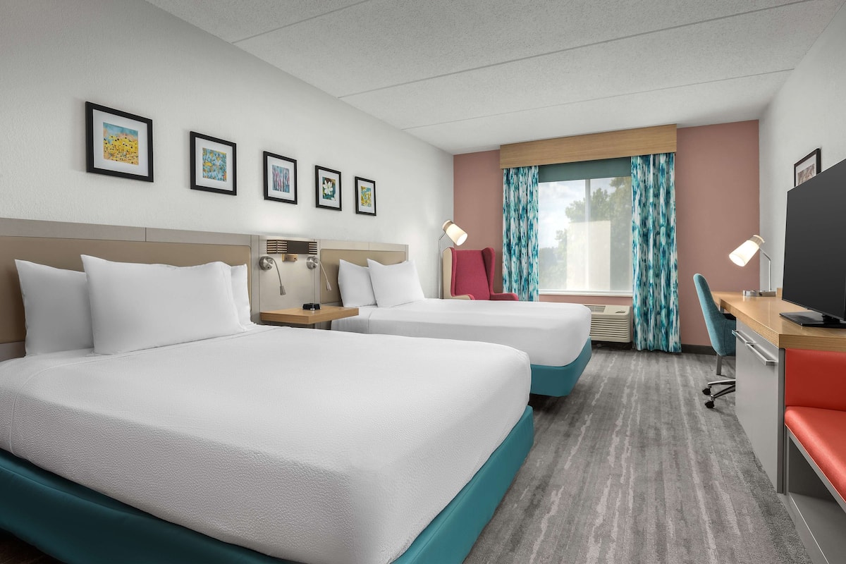 A hotel room with two beds, a desk, a TV, and a window with curtains.