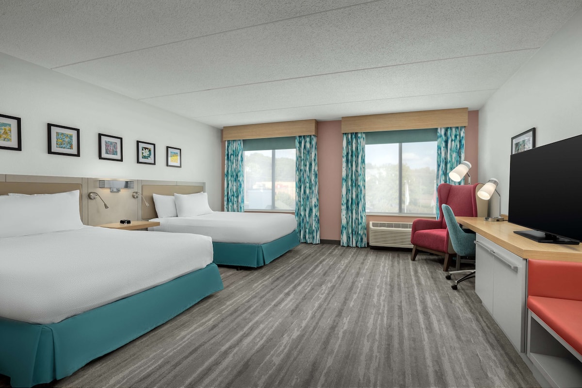 A hotel room with two beds, a desk with a TV, a chair, and a window with curtains.