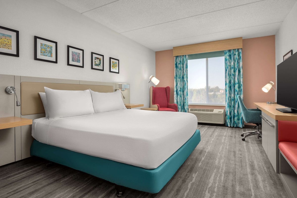 A hotel room with a large bed, a desk, a chair, a TV, and a window with curtains.