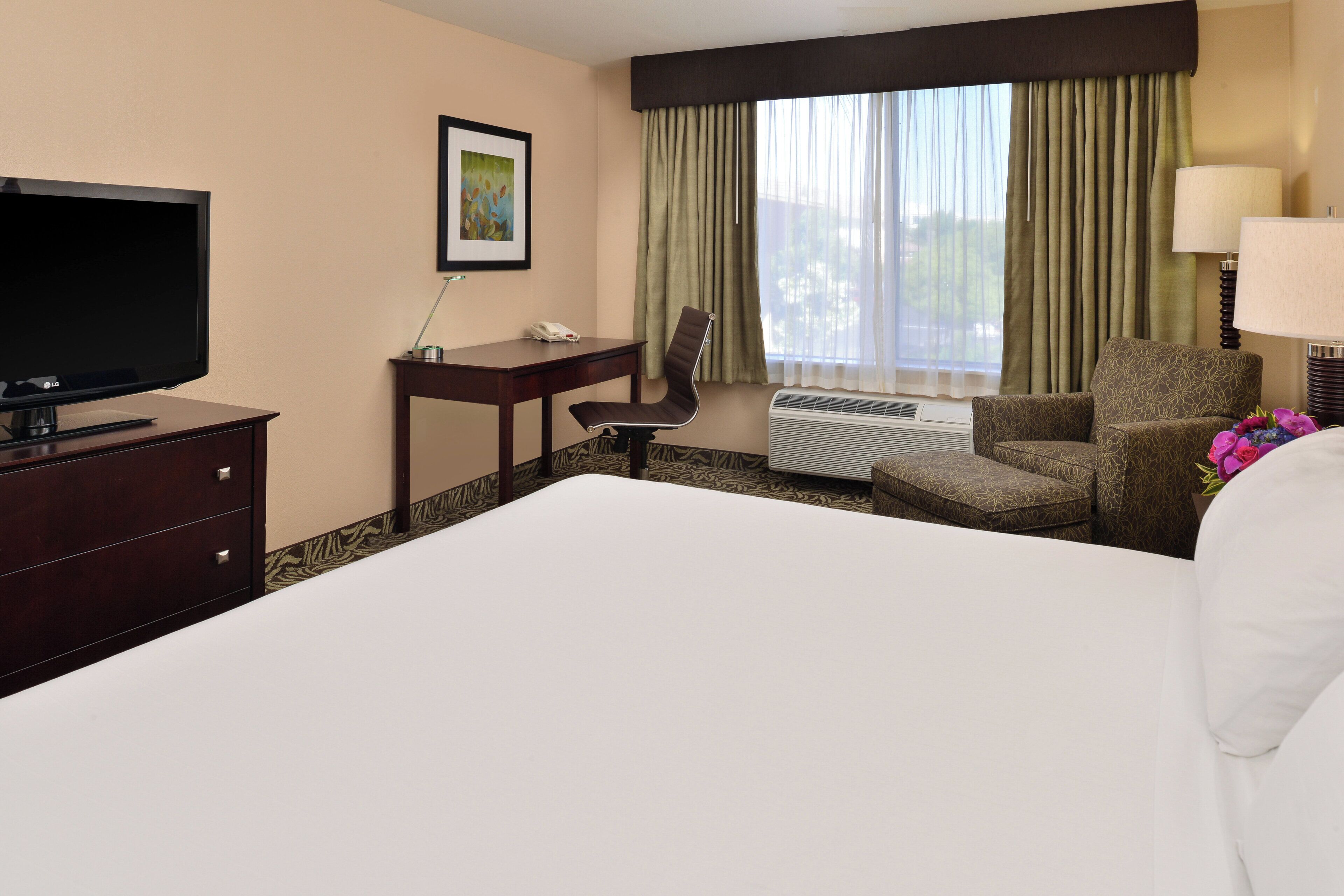 Une chambre d’hôtel avec un grand lit, un bureau avec un téléphone, une chaise, une télévision et une fenêtre avec des rideaux.