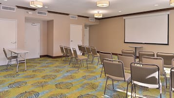 Un salón de conferencias con filas de sillas, una pantalla de proyector y una alfombra estampada con motivos de hojas.
