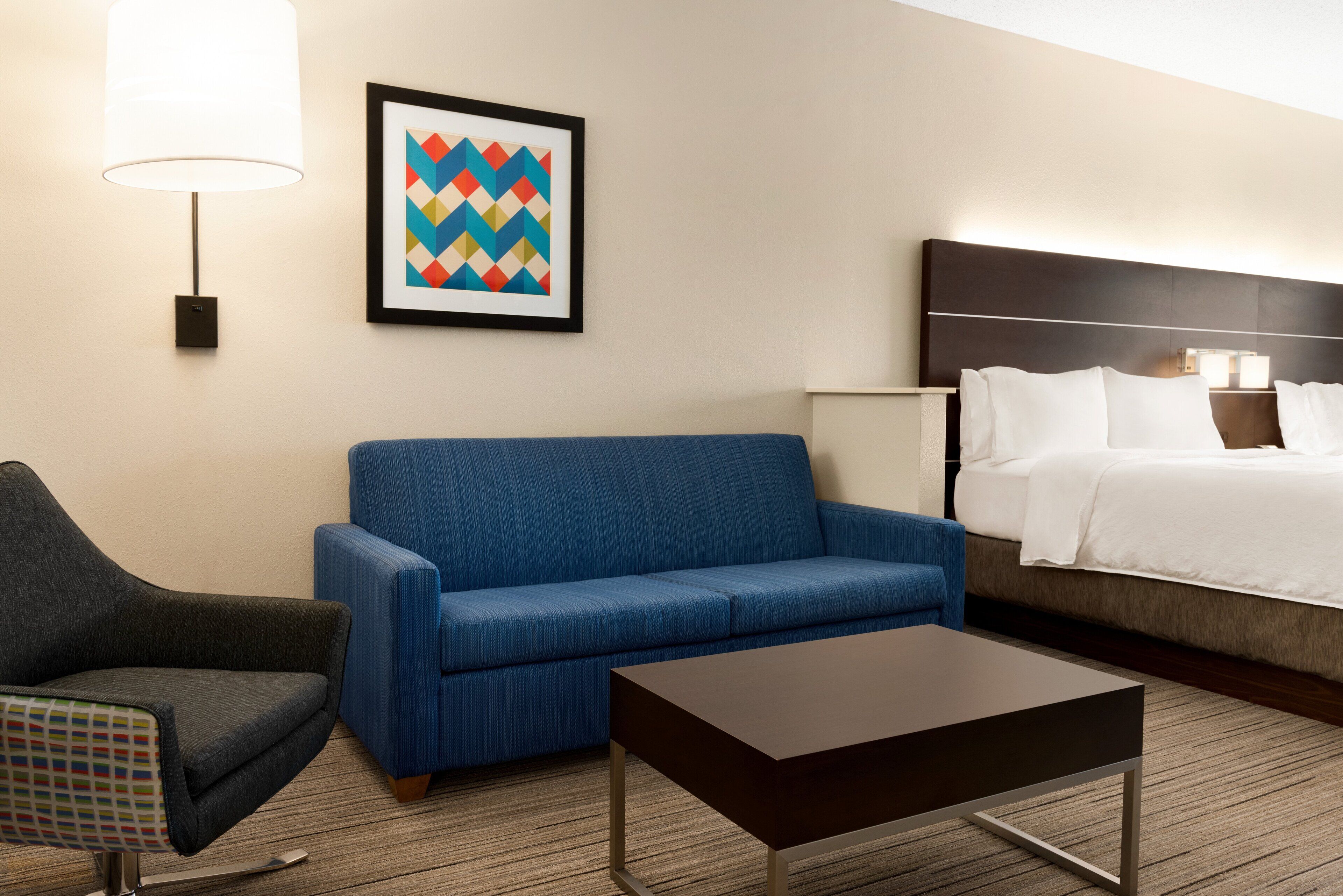 A hotel room with a blue sofa, a dark armchair, a small table, a bed with white linens, a framed geometric artwork, and a wall-mounted lamp.