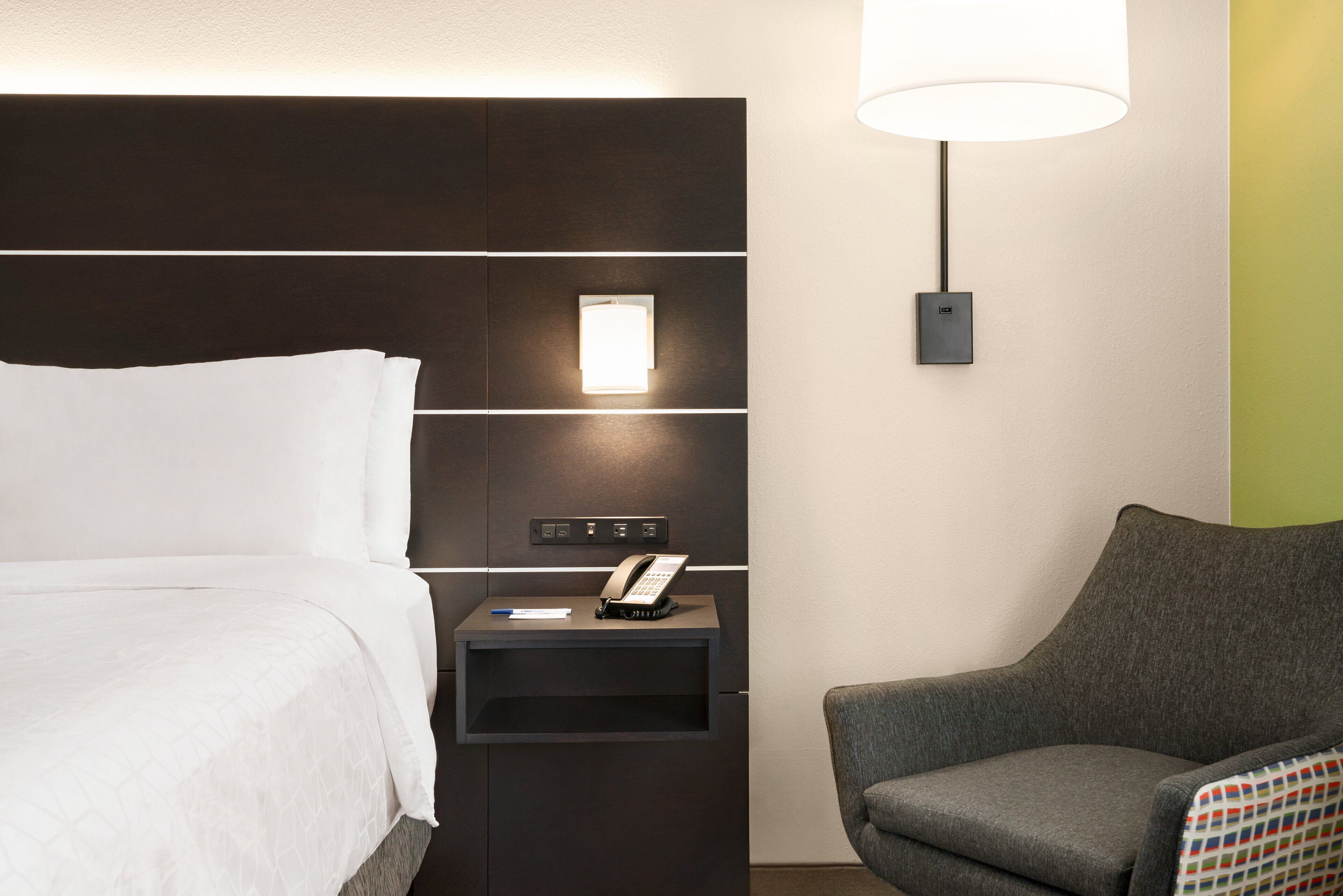 A hotel room with a bed, a bedside table with a phone, a lamp, and a chair.