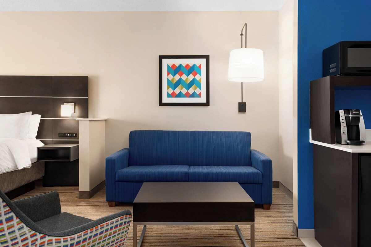 A modern hotel room with a blue sofa, a small coffee table, a chair, a bed with white linens, a bedside table with a lamp, and a framed artwork on the wall.