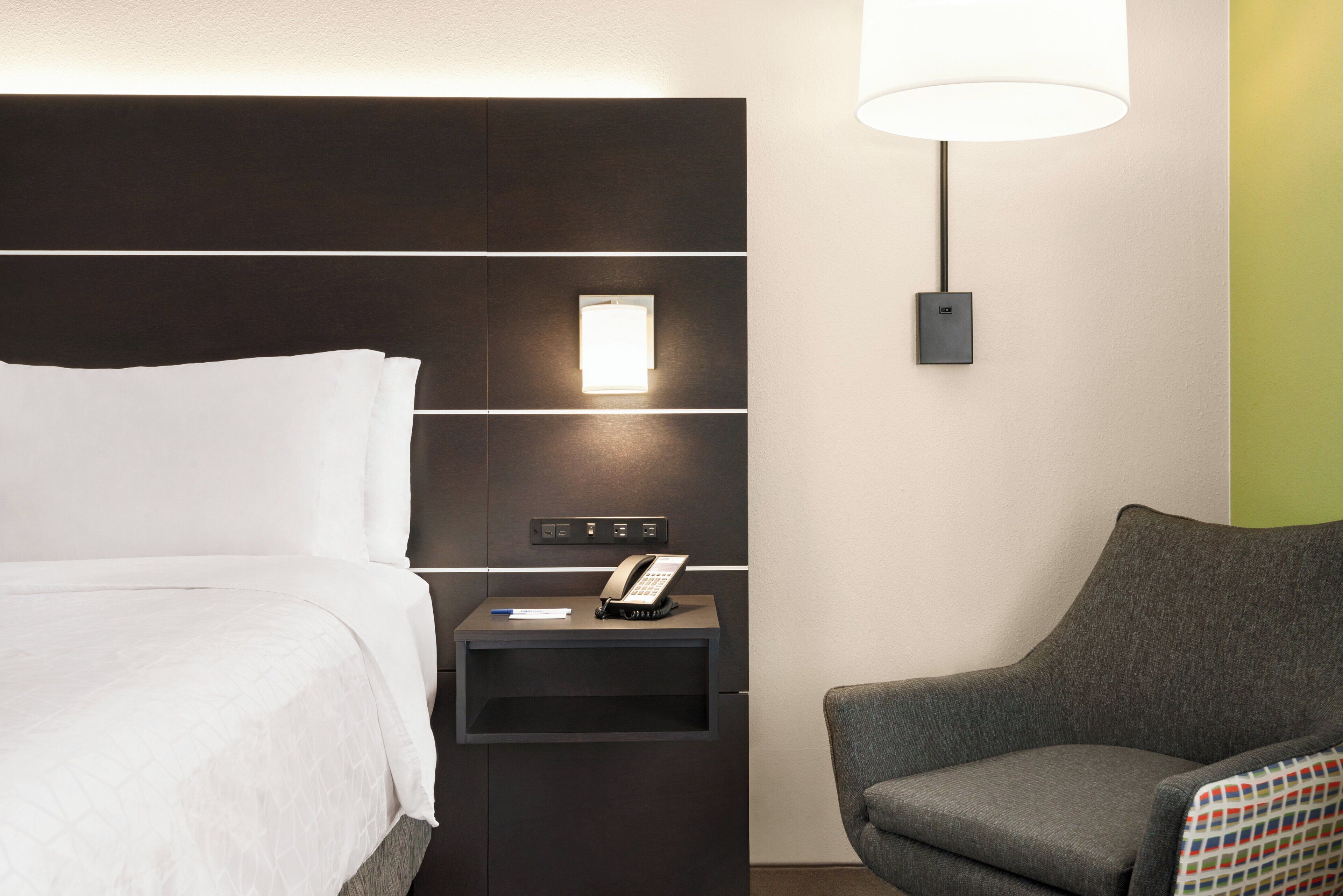 A hotel room with a bed, a bedside table with a phone, a lamp, and a chair.