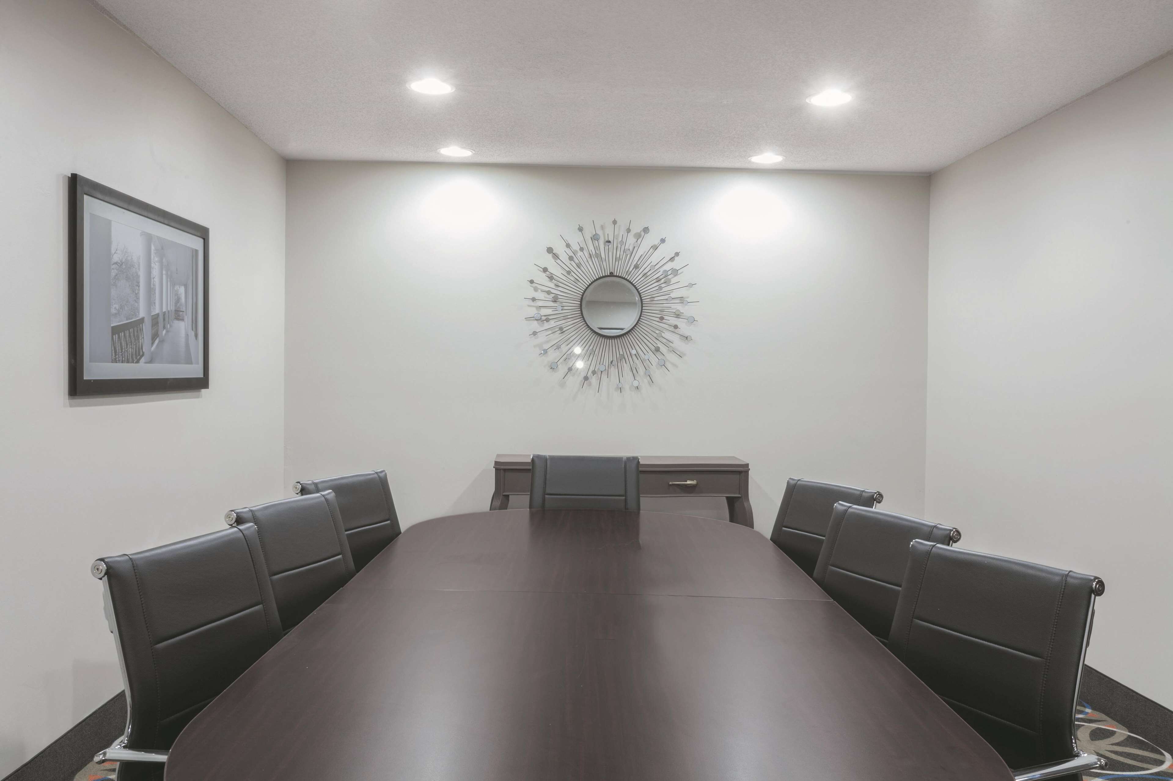 A conference room with a long table, six chairs, a small desk, a framed picture on the wall, and a decorative mirror.