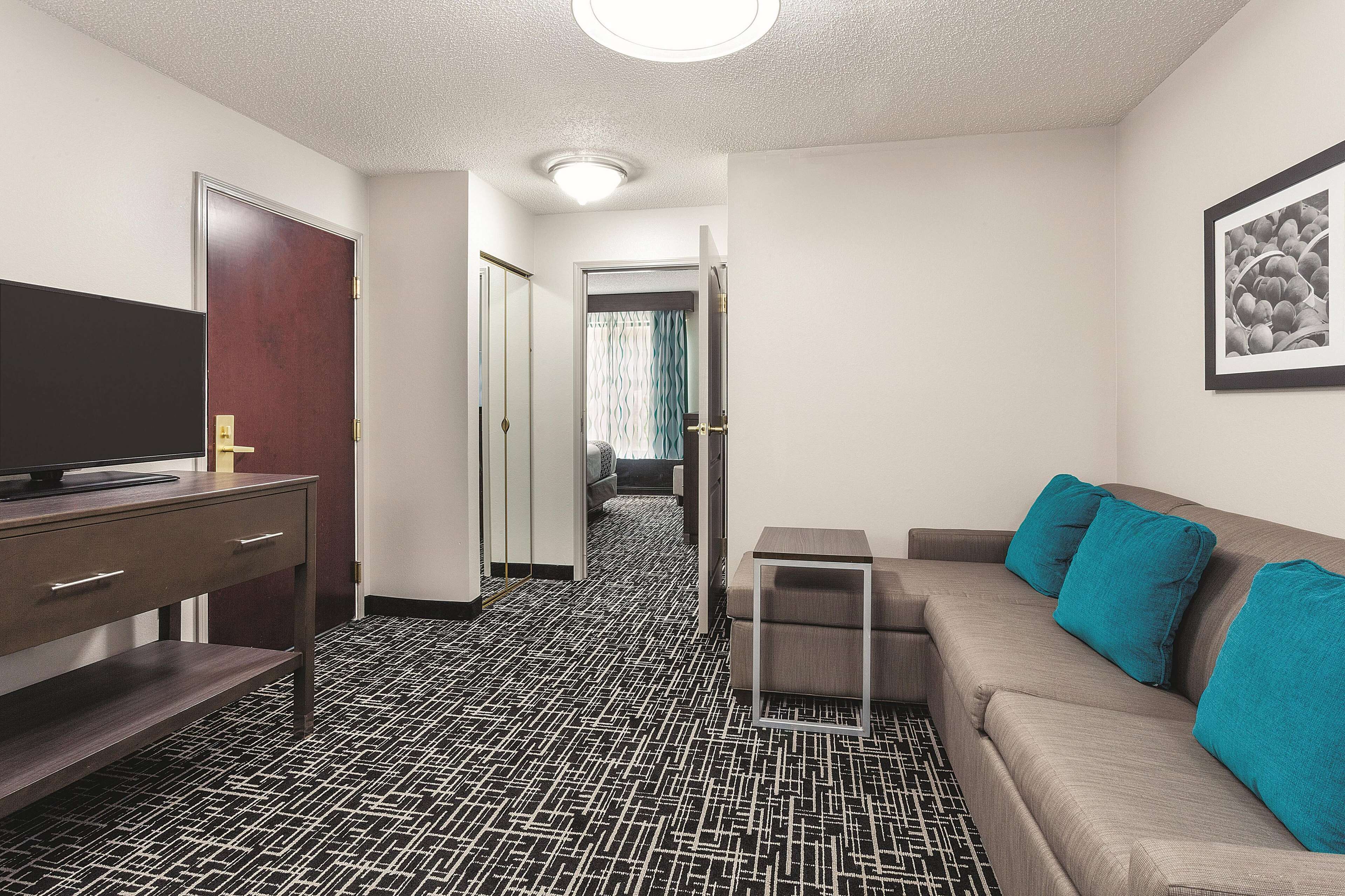 A hotel room with a black and white patterned floor, a sofa with teal cushions, a TV on a wooden stand, and a framed picture on the wall.