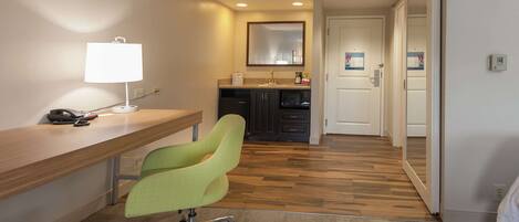 A hotel room with a wooden desk, a green office chair, a desk lamp, a microwave, and a small kitchenette.