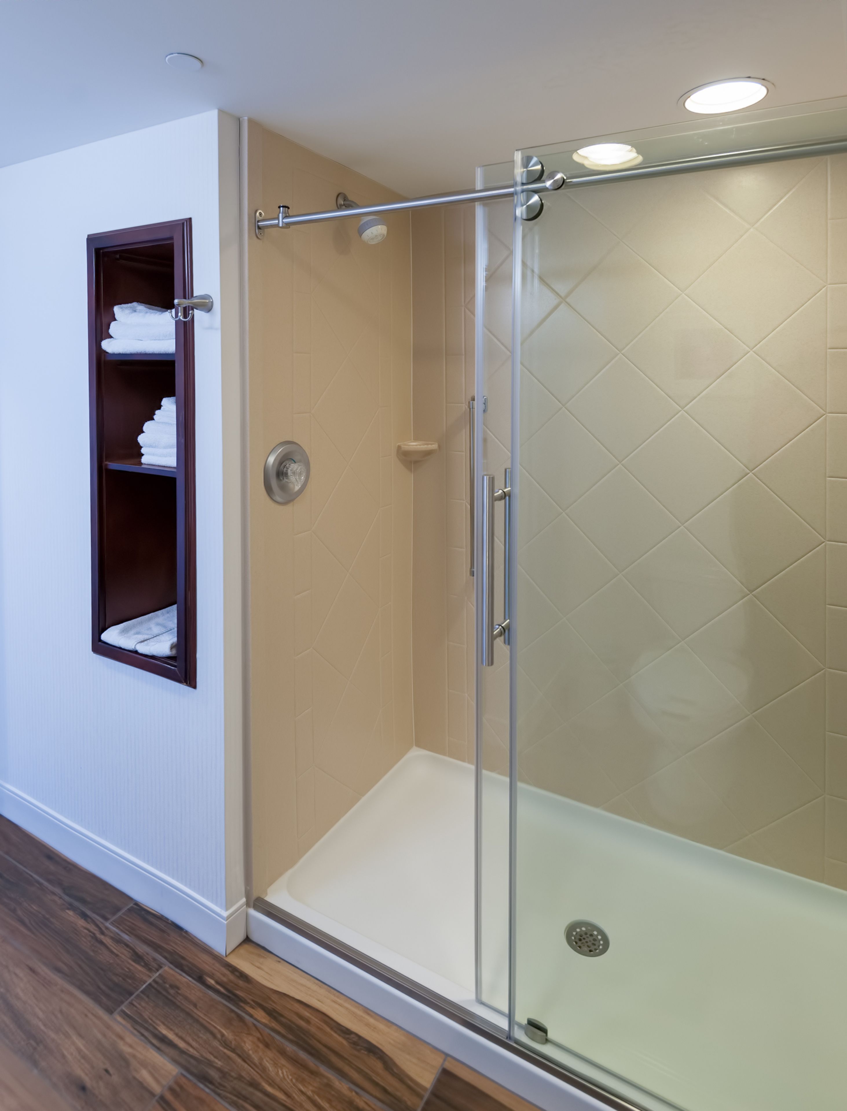 Una ducha con mampara de vidrio, pared con azulejos, estante de madera con toallas y suelo de madera.
