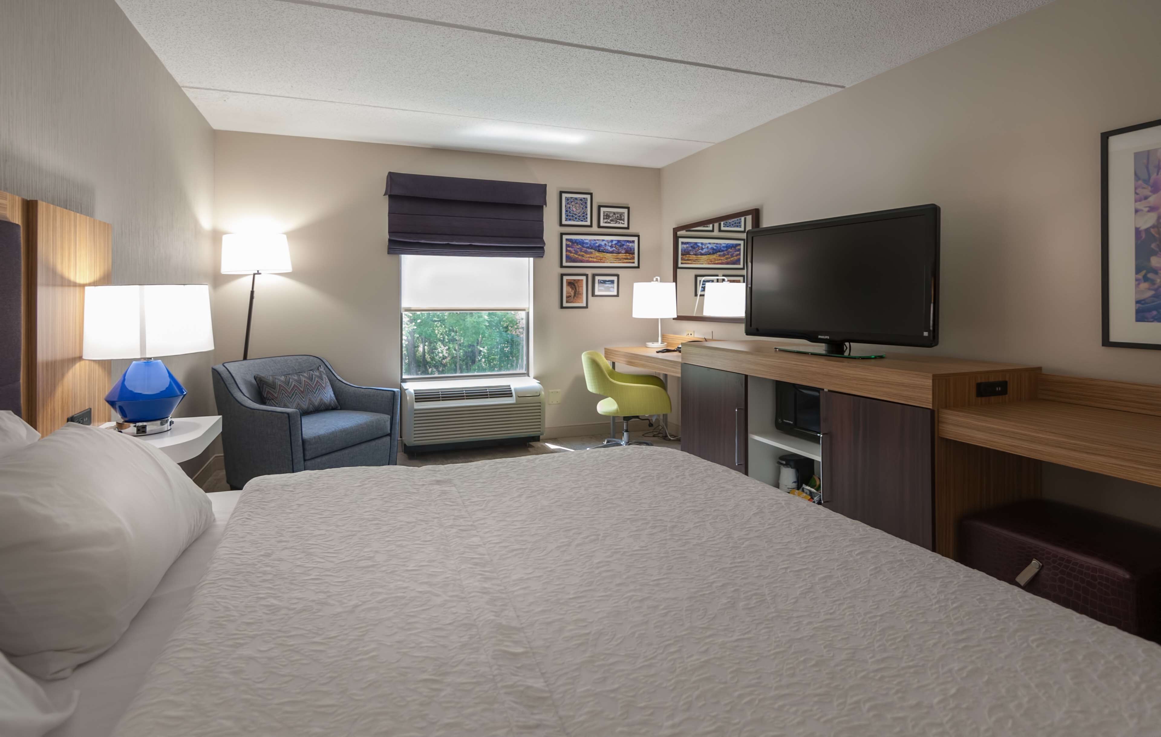 A hotel room with a large bed, a desk with a flat-screen TV, a chair, a lamp, and a window with a view of greenery.