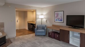Ein modernes Hotelzimmer mit Holzboden, einem blauen Sessel, einem Fernseher, einem Schreibtisch und einem durch eine offene Tür einsehbaren Badezimmer.