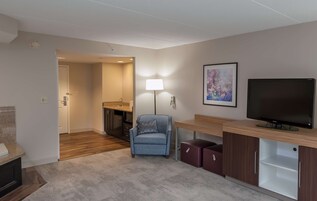 Quarto de hotel moderno com piso de madeira, uma poltrona azul, televisão, escrivaninha e banheiro visível através de uma porta aberta.