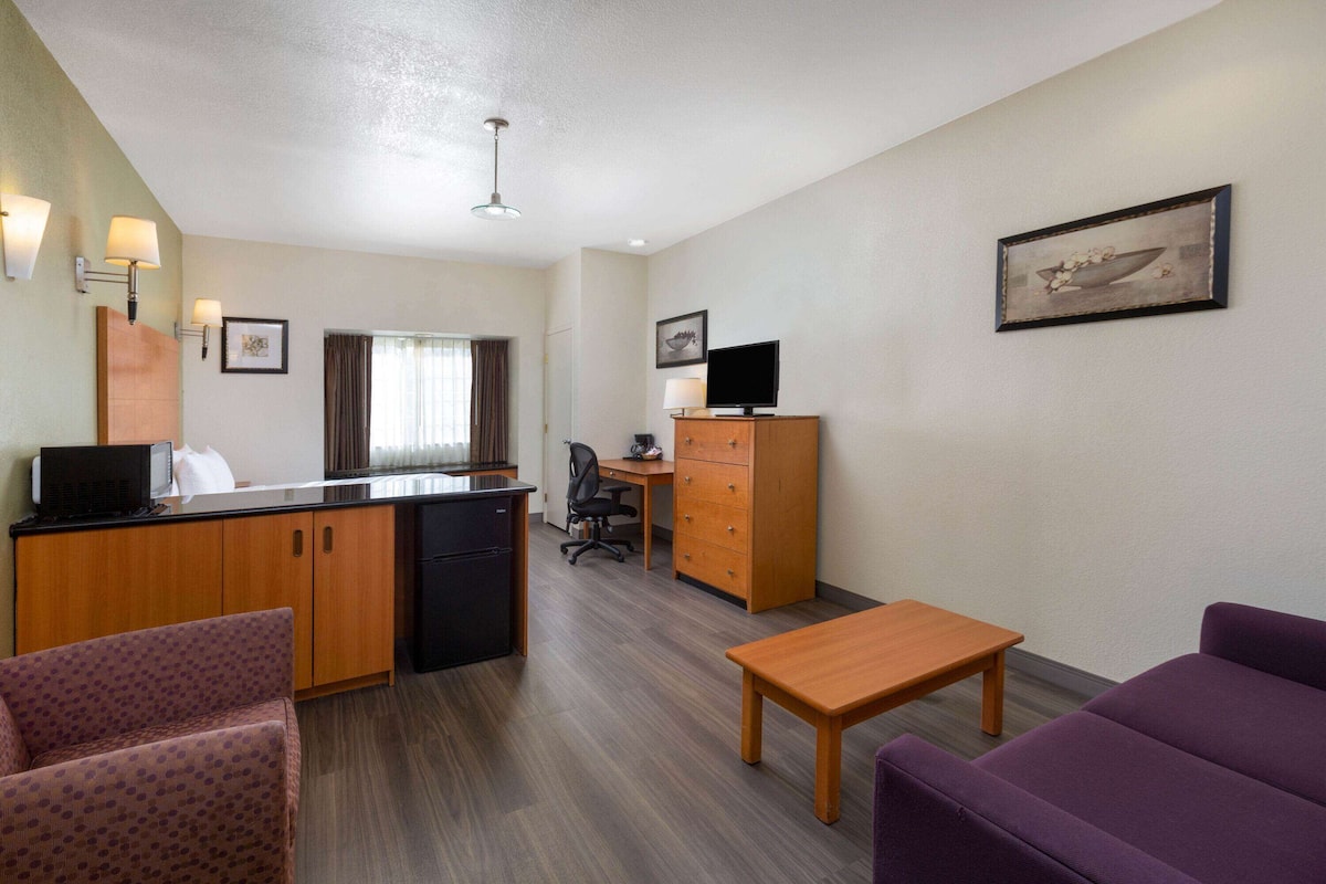 A hotel room with a kitchenette, a desk, a chair, a sofa, and a television.