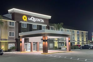 A La Quinta hotel with a modern design, featuring a prominent sun logo, a covered entrance, and a parking lot with several vehicles.