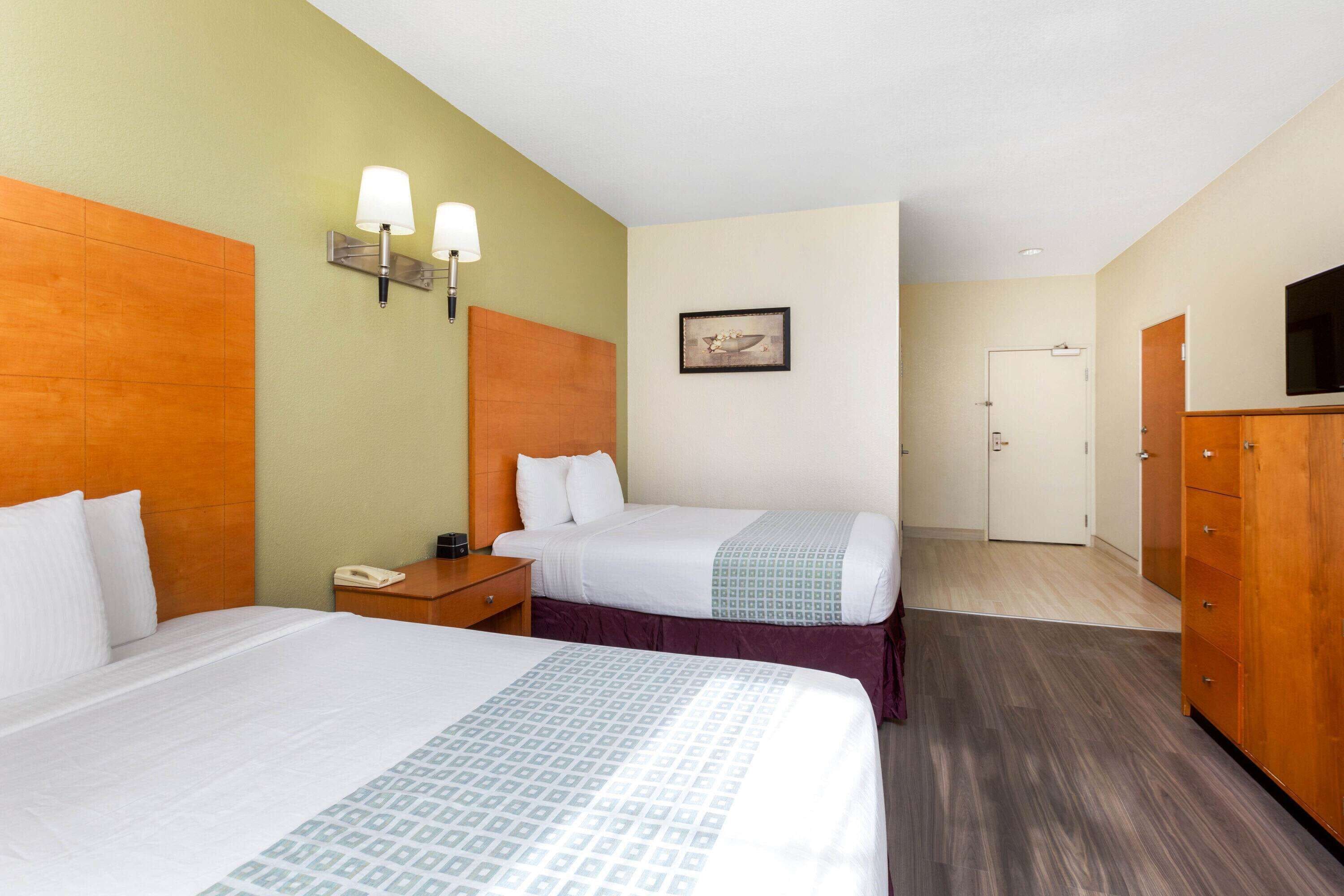 Une chambre d’hôtel avec deux lits, une table de chevet, un bureau, une chaise, une armoire, un téléviseur et un tableau encadré au mur.