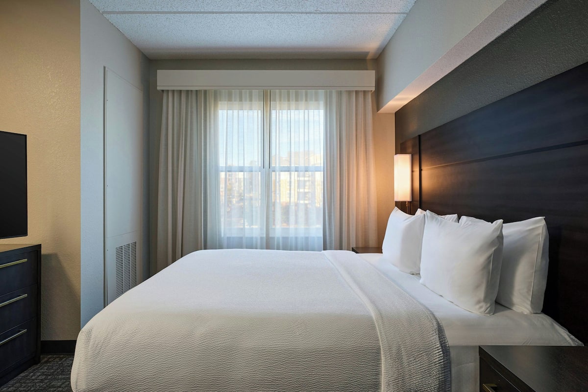 A hotel room with a large bed, a flat-screen TV, a dresser, and a window with curtains.