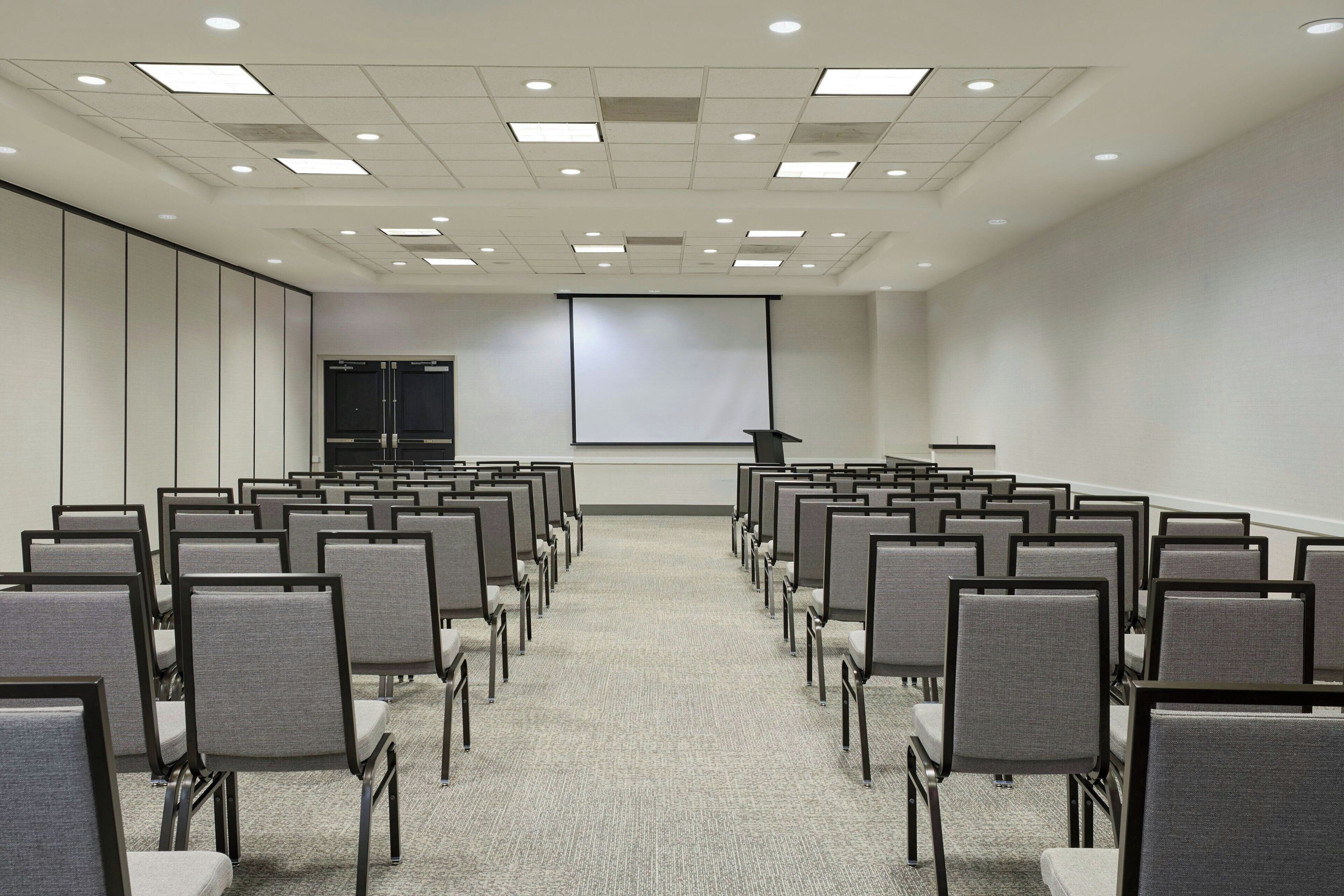 Uma sala de conferências com fileiras de cadeiras, uma tela de projetor e piso carpeteado.