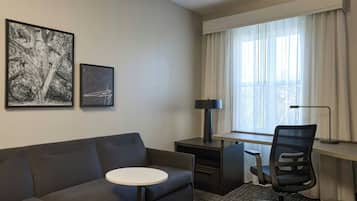 A hotel room with a gray sofa, a small round table, a desk with a chair, a lamp, and two framed pictures on the wall.