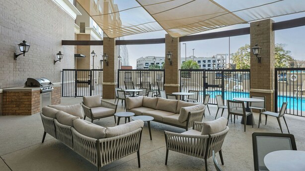A covered outdoor seating area with wicker furniture, a barbecue grill, and a view of a swimming pool and buildings in the background. - Residence Inn Tallahassee Universities at the Capitol (Tallahassee)