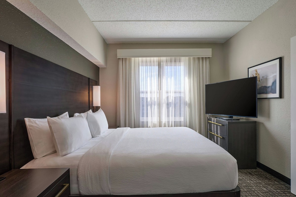 A hotel room with a large bed, a flat-screen TV, a desk with drawers, and a window with sheer curtains.