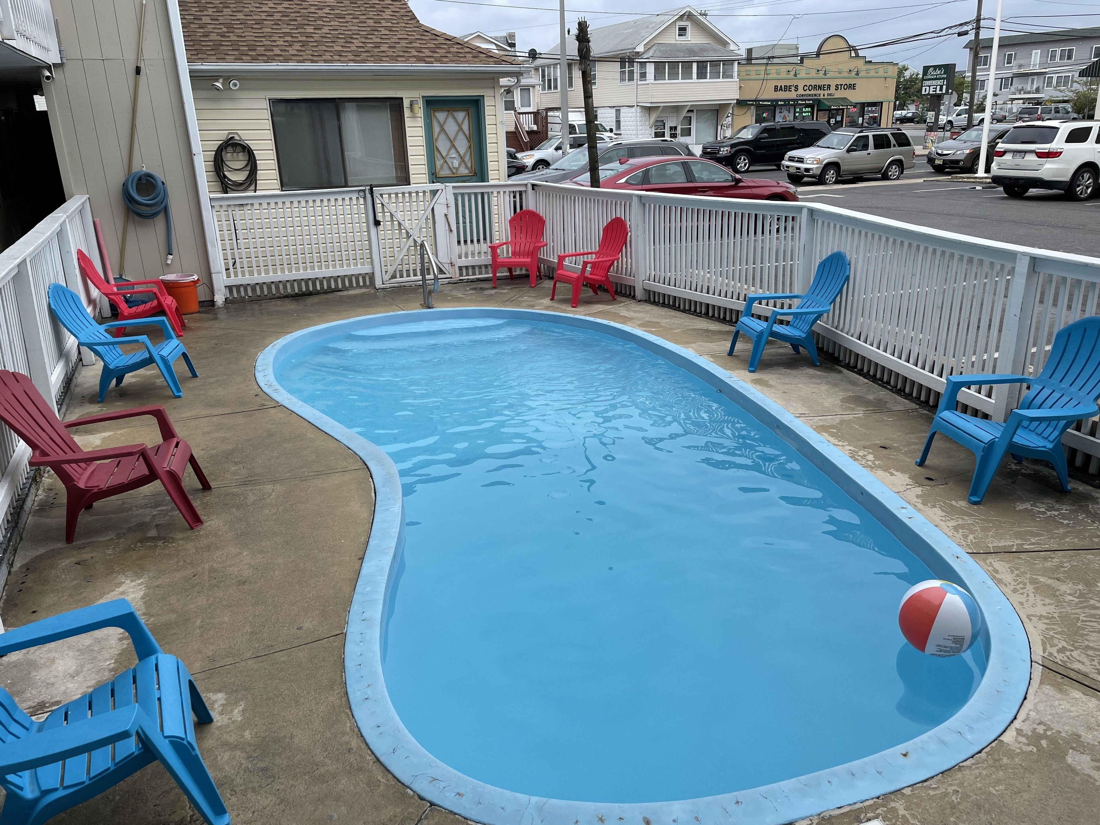 A swimming pool with blue and red Adirondack chairs around it, a beach ball, and a white picket fence.