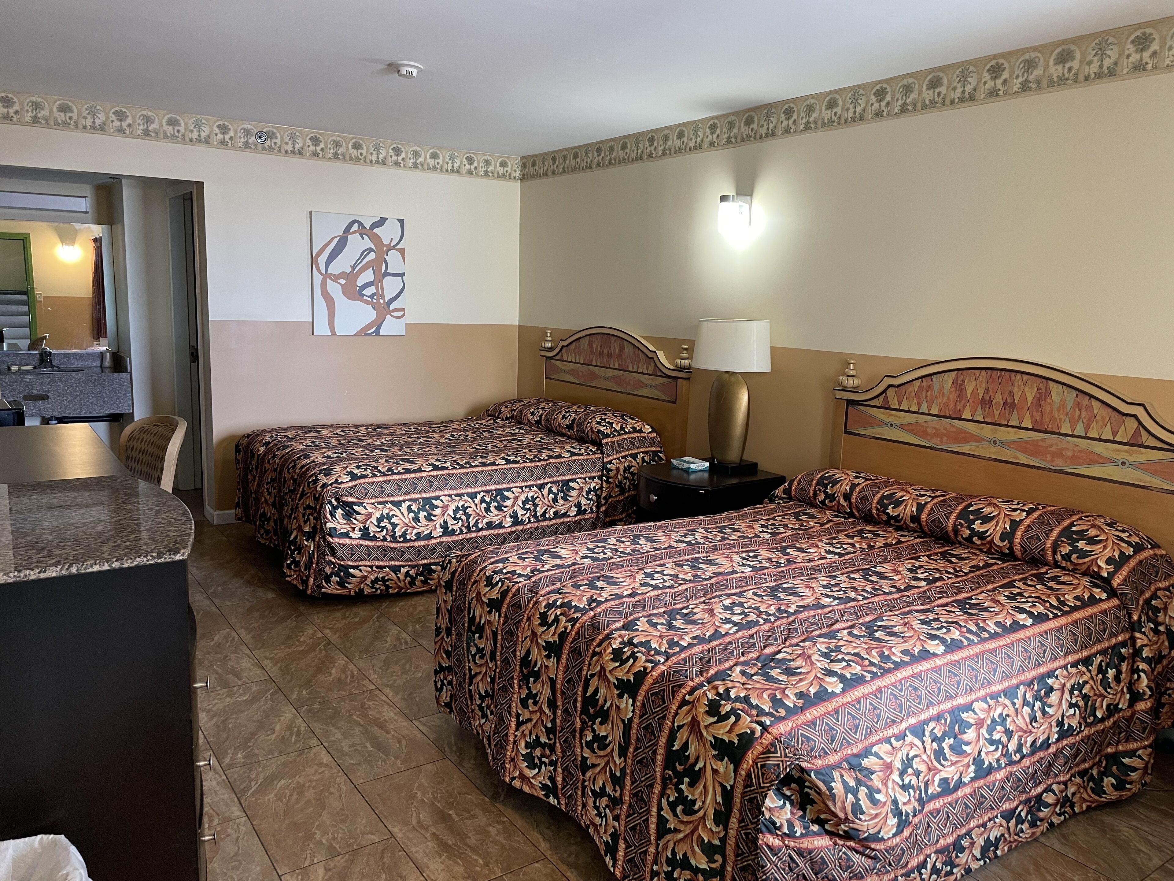 A hotel room with two beds, a desk, a chair, a lamp, and a bathroom visible through an open door.