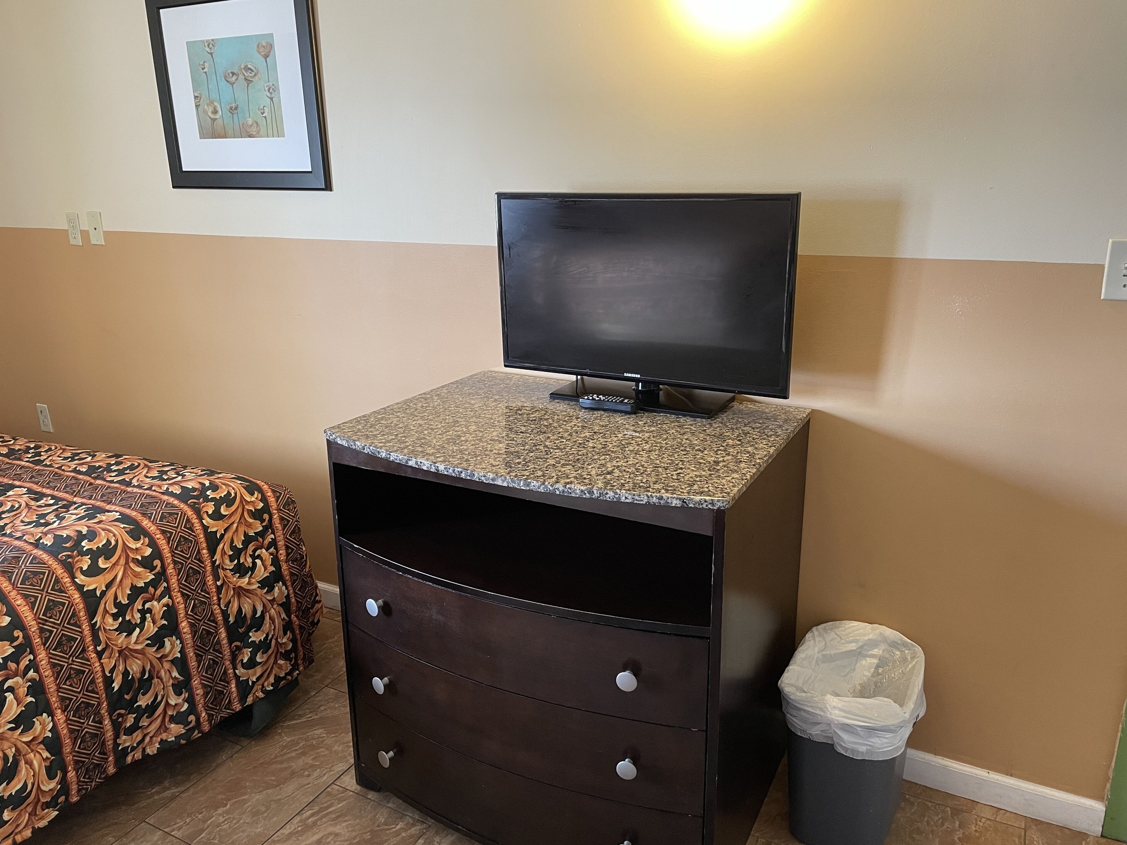 A small room with a bed, a TV on a granite counter, a trash bin, and a framed picture on the wall.