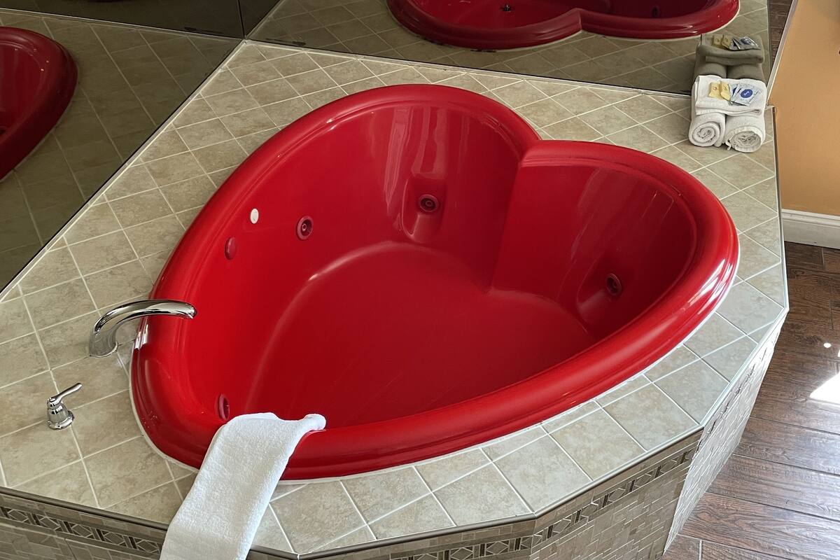 A heart-shaped red bathtub with a white towel, surrounded by beige tiles and a patterned border.