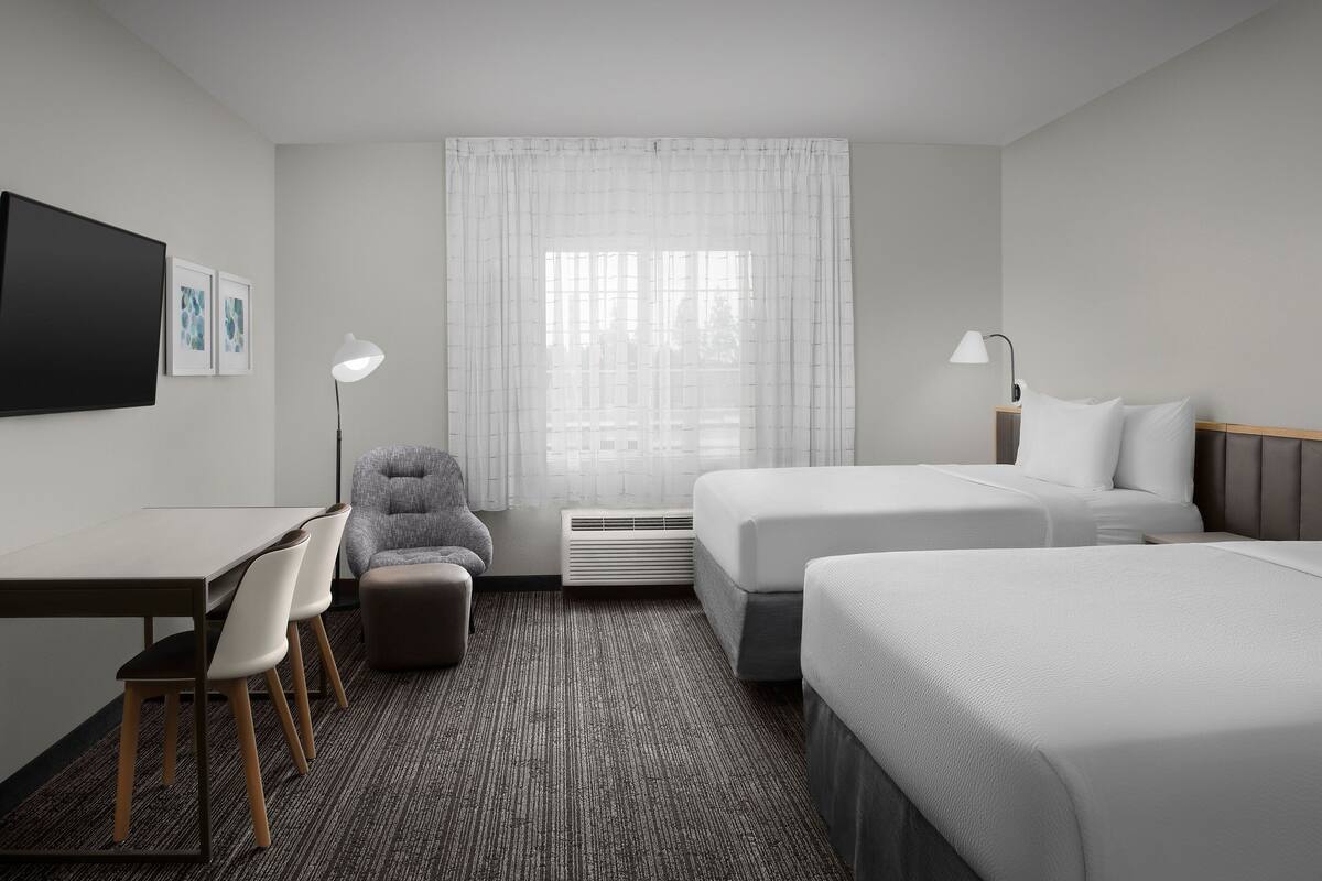 Una habitación de hotel con dos camas, un escritorio con sillas, un televisor y una ventana con cortinas.