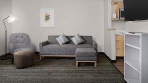 Ein modernes Wohnzimmer mit grauem Sofa, passendem Hocker und Stehlampe. Ein gerahmtes Kunstwerk ist an der Wand, und ein Flachbildfernseher ist über einer Küchenzeile angebracht.