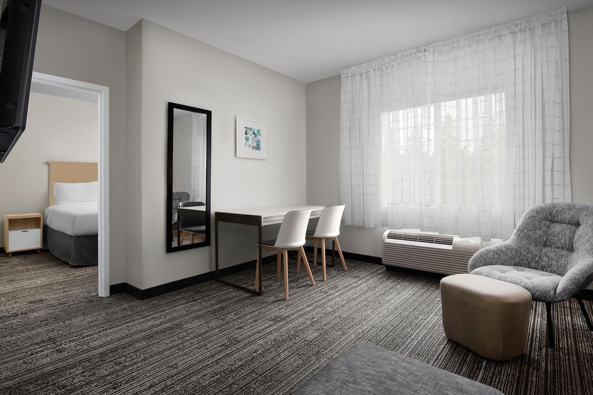 Habitación de hotel con cama, escritorio con sillas, espejo grande y ventana con cortinas translúcidas.