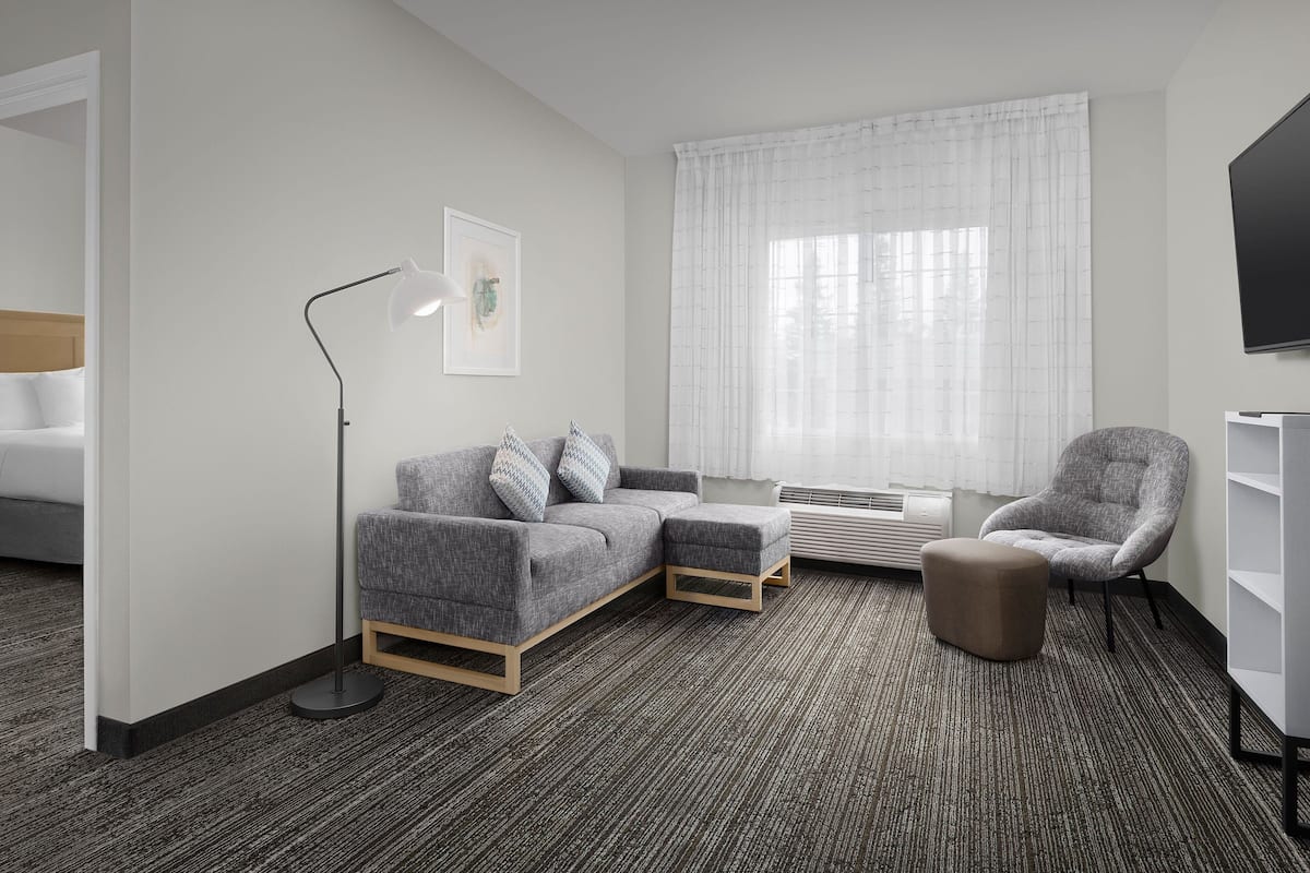 Una habitación de hotel moderna con un sofá gris, un sillón a juego, una lámpara de pie, un televisor de pantalla plana y un ventanal con cortinas translúcidas.