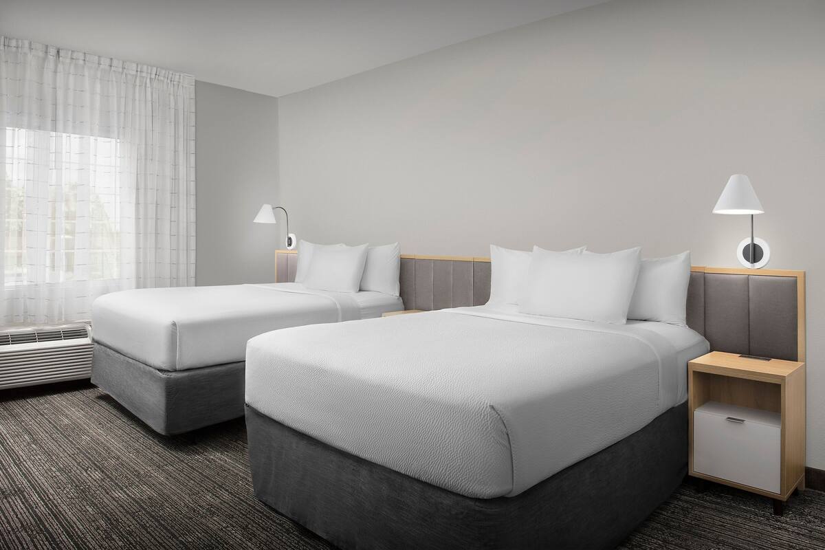 Habitación de hotel con dos camas, una mesita de noche con lámpara, un calentador y una ventana con cortinas.