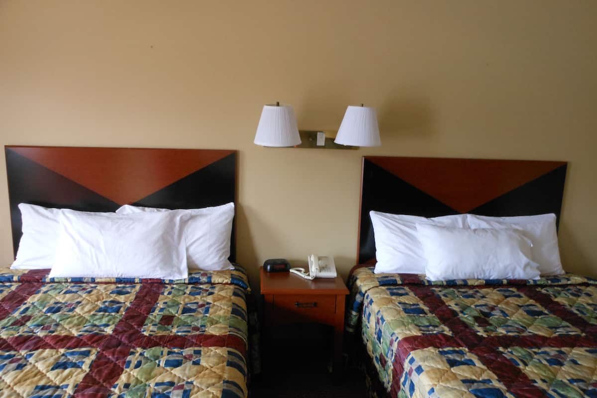 Habitación de hotel con dos camas, cada una con cubrecama acolchado y almohadas blancas. Hay dos lámparas de noche y un teléfono en la mesita entre las camas.