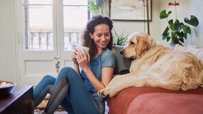 A person sitting on a bed with a dog, holding a cup, in a room with a door and a plant. - Staybridge Suites Davenport by IHG (Davenport)