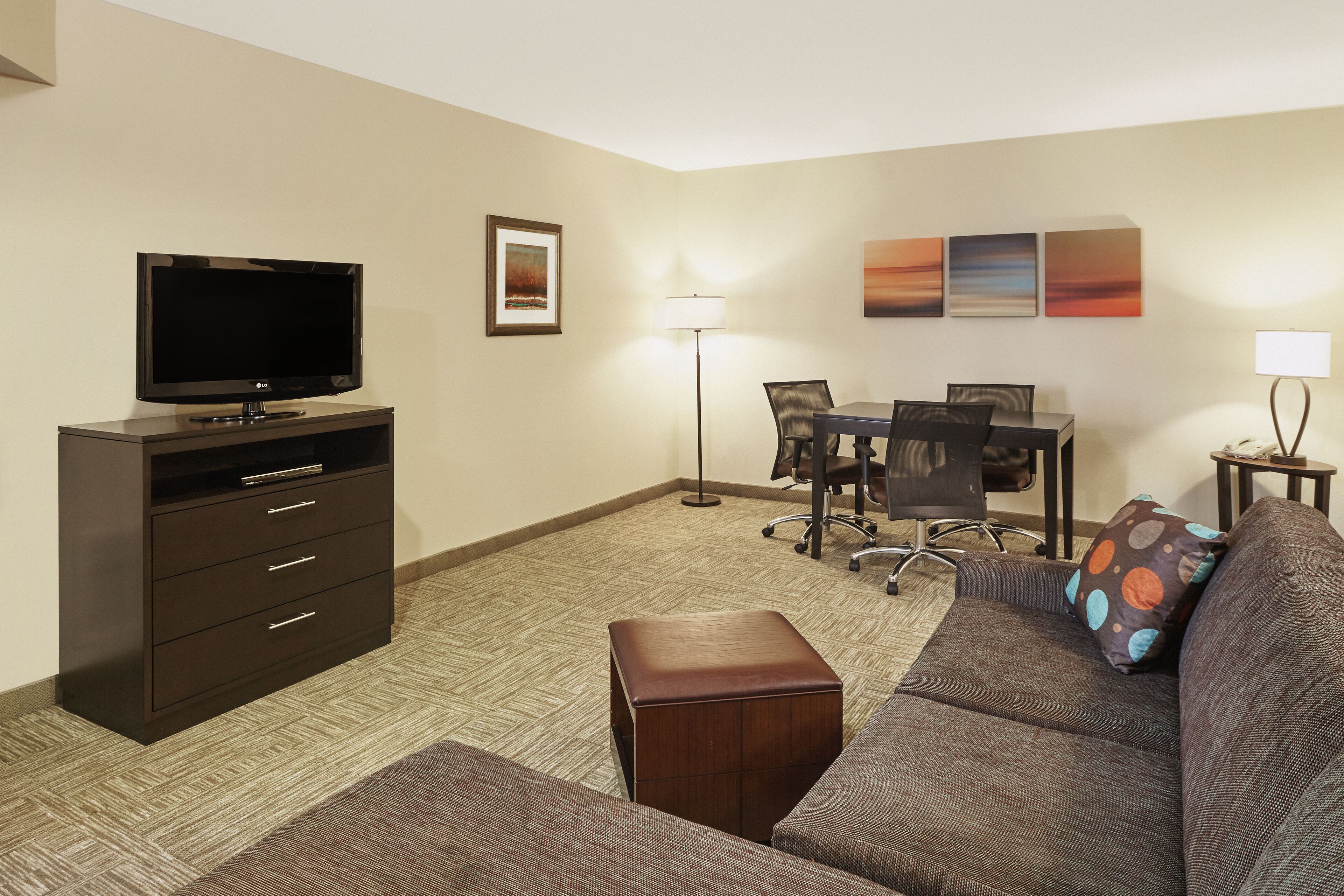 A hotel room with a flat-screen TV, a desk with a chair, a sofa with a pillow, a lamp, and artwork on the wall.